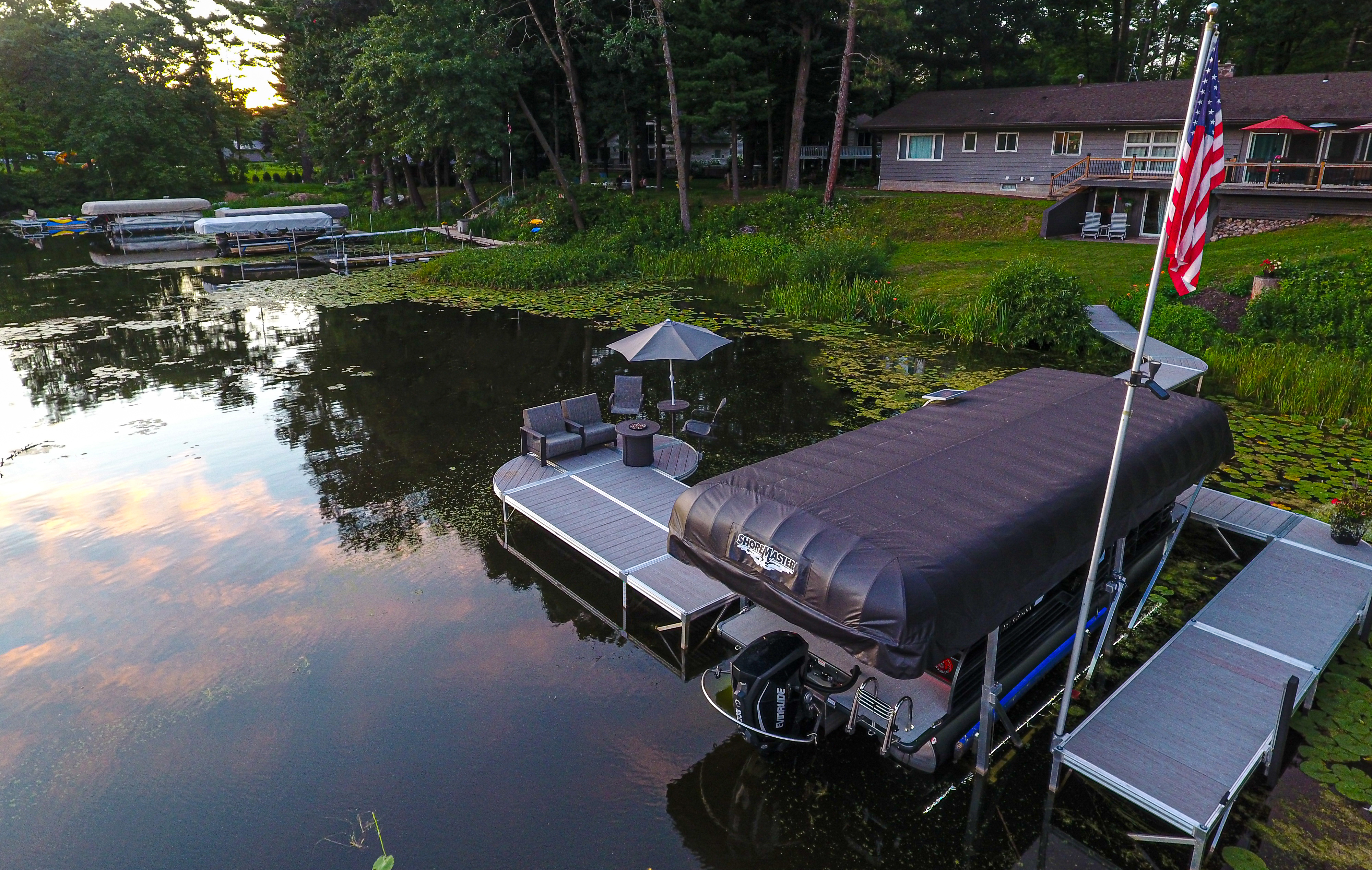 An aerial view of a waterfront property featuring a modular dock system, a boat lift with a black canopy cover and outdoor seating with an umbrella.